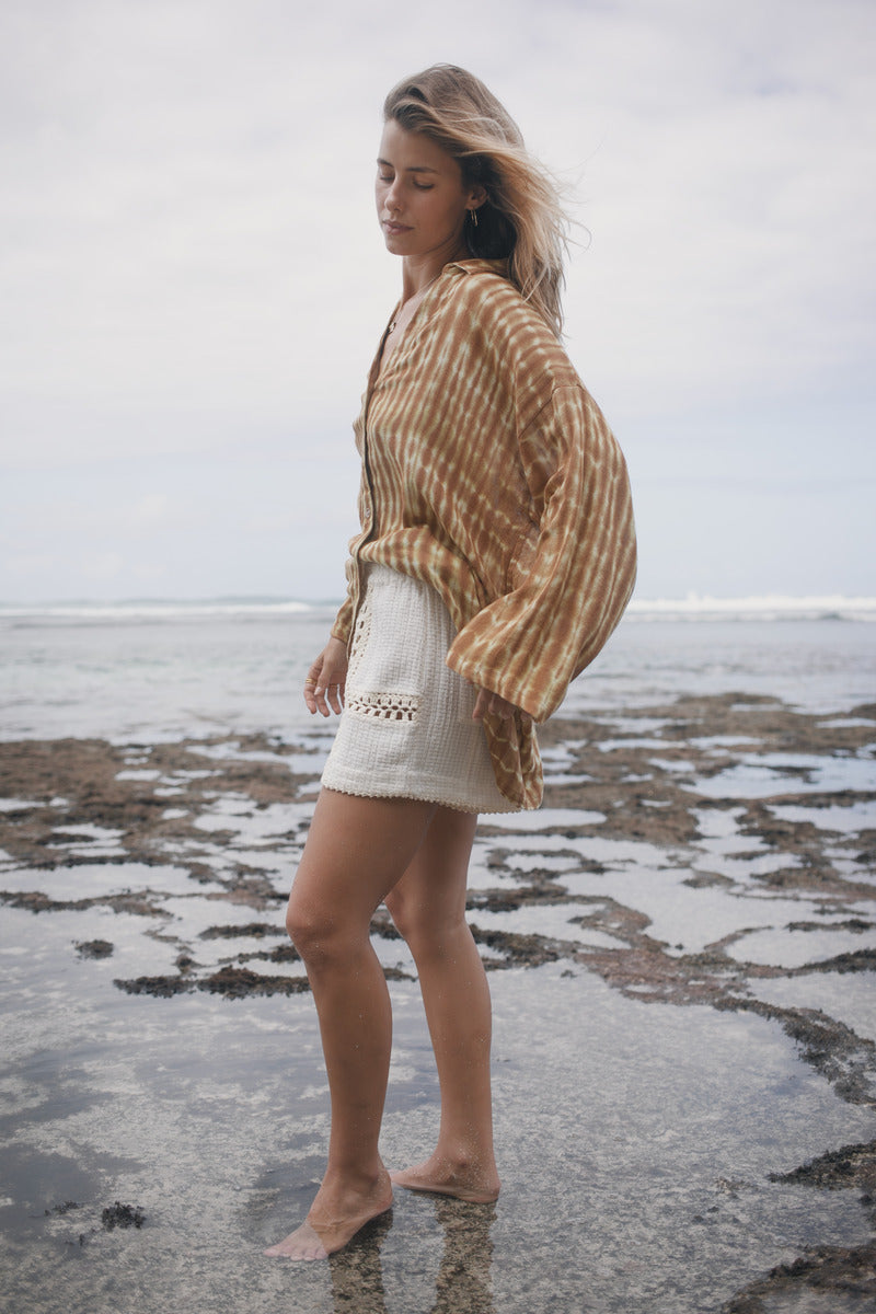 Woman standing on a rocky beach wearing a patterned shawl and white shorts.