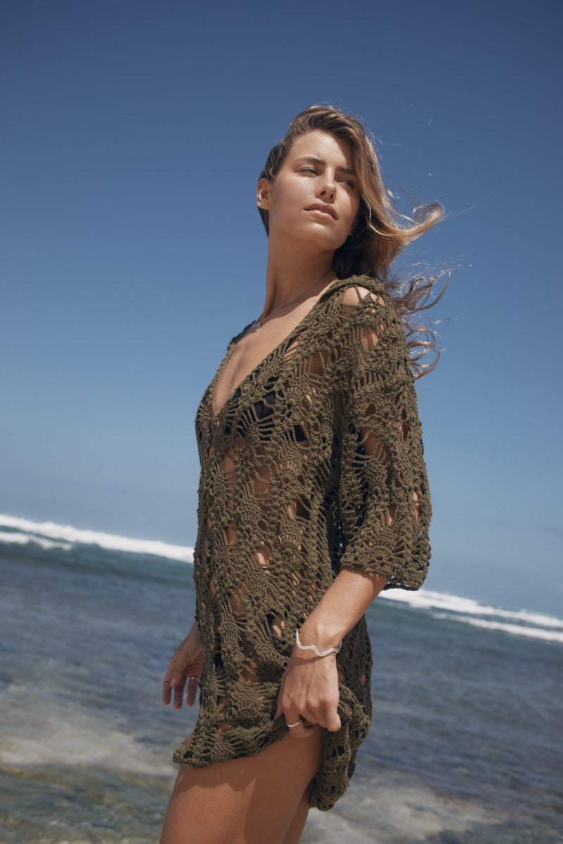 Woman in a crochet cover-up standing on a beach with waves in the background