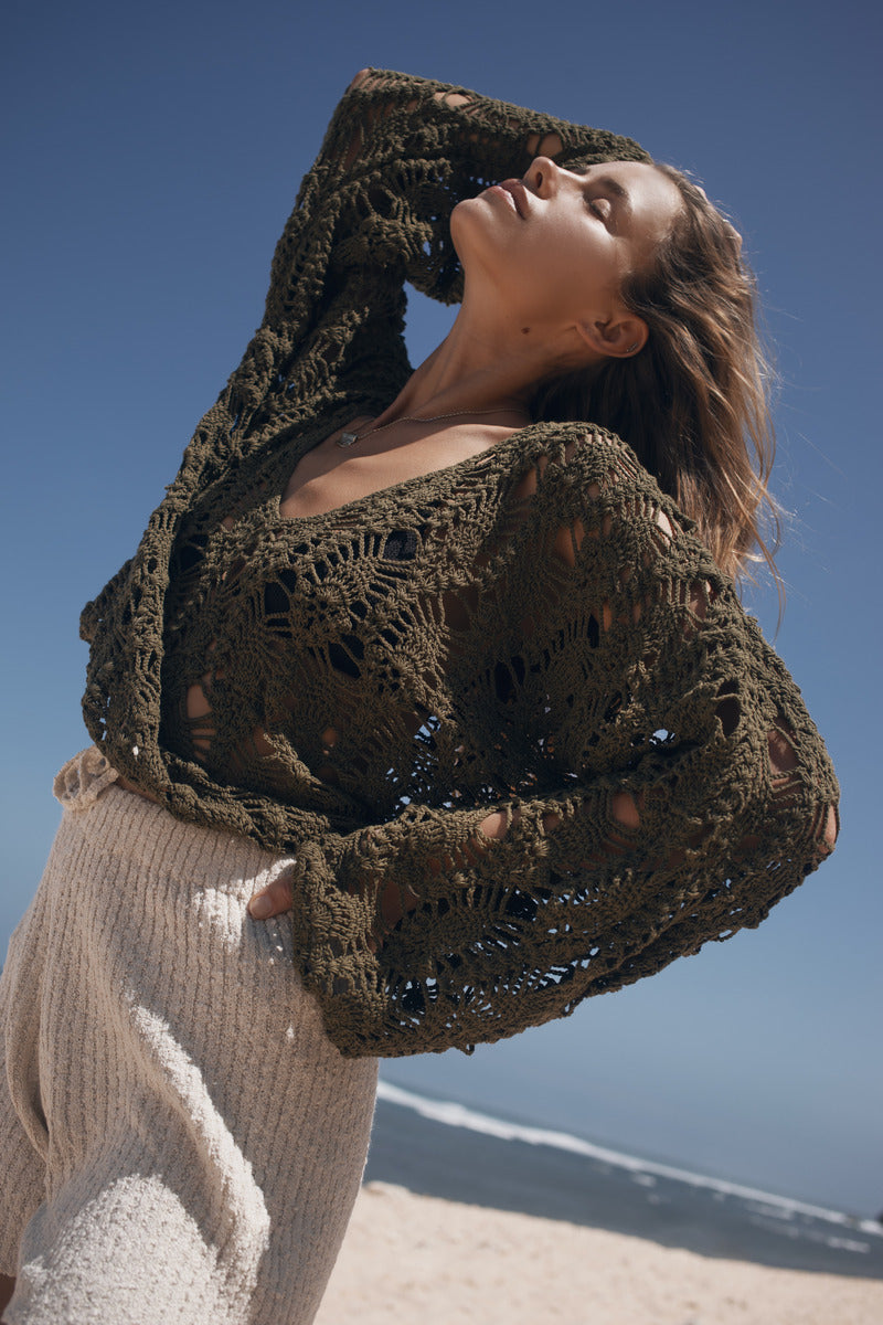 Woman wearing a large green knitted crochet top on a beach with clear blue sky.