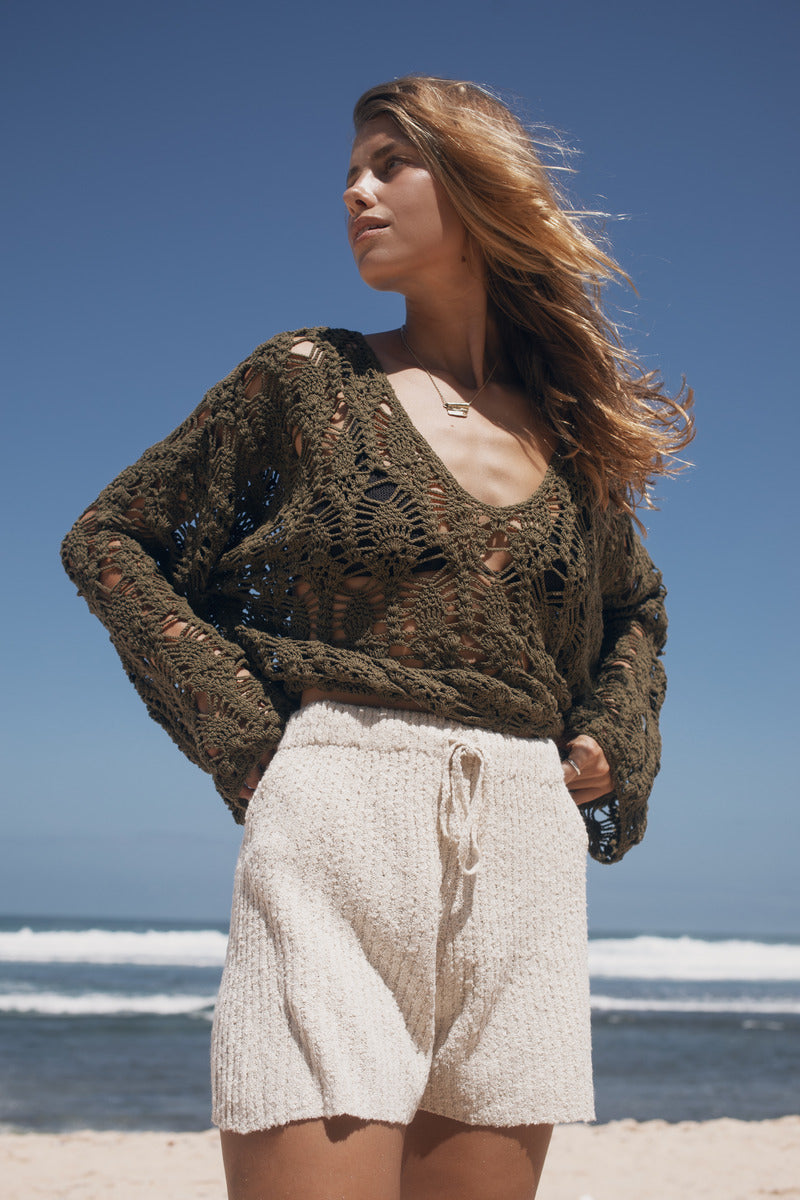 Woman wearing a green crochet sweater and beige shorts standing on a beach.