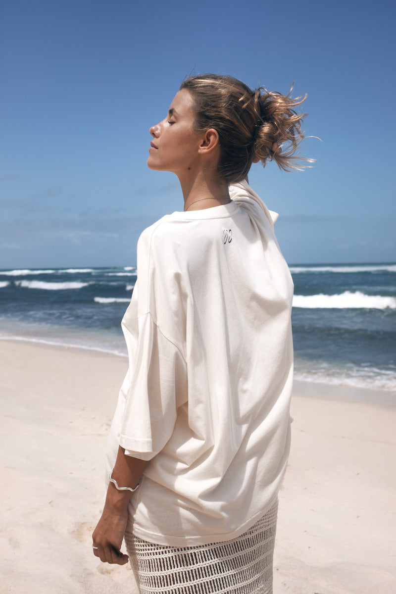 Woman in a white outfit standing on a beach with waves and clear sky.