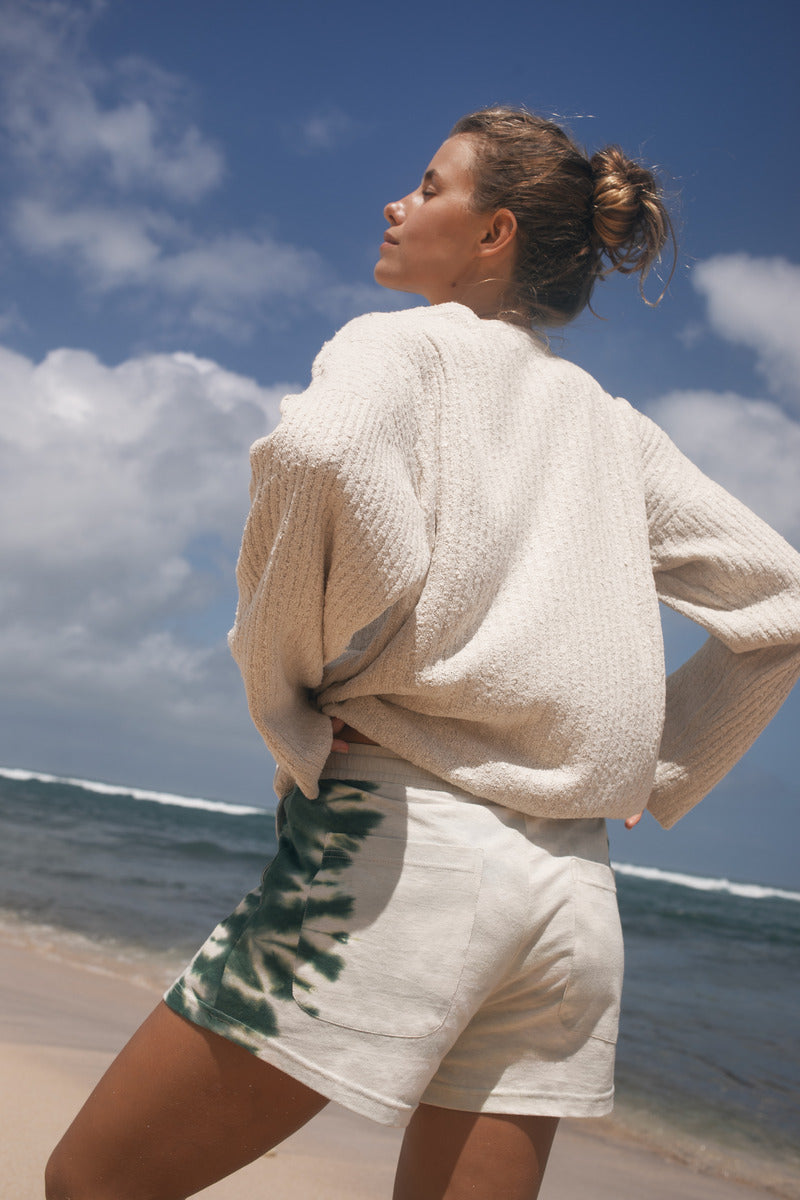 Woman in a beige cardigan and white shorts standing on a beach with ocean and sky in the background