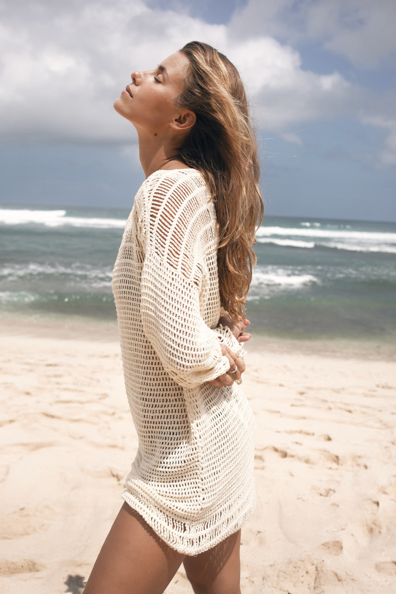 Woman in a white crochet mini dress on a beach with ocean waves in the background