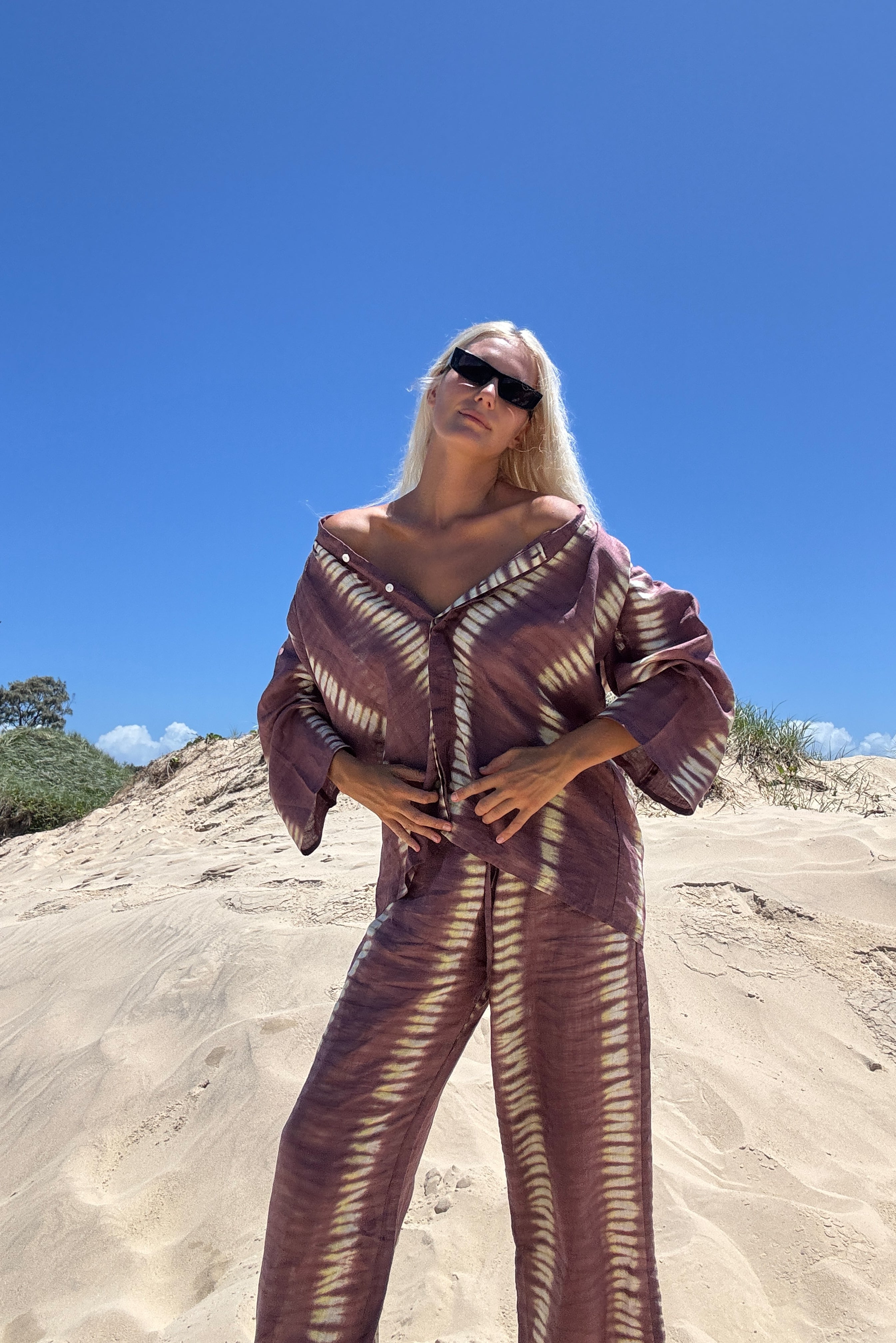 A person standing on a sandy surface wearing a deep mauve tie-dye shirt with wide sleeves and a V-neckline, paired with matching pants, sunglasses, and a handbag.