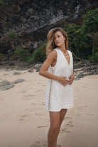 Woman in a white dress standing on a beach with rocky and greenery background