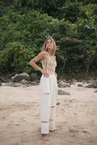 Woman in a striped top and white pants standing on a beach with greenery in the background