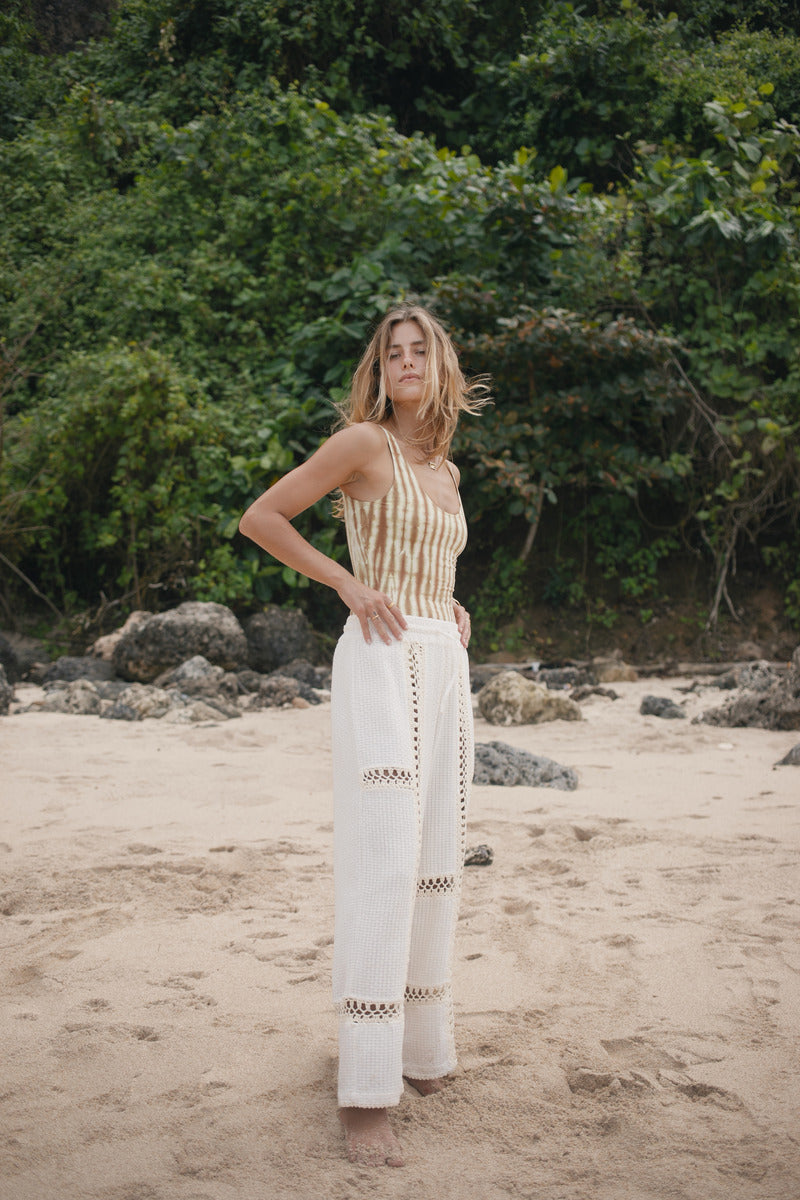 Woman in a striped top and white pants standing on a beach with greenery in the background