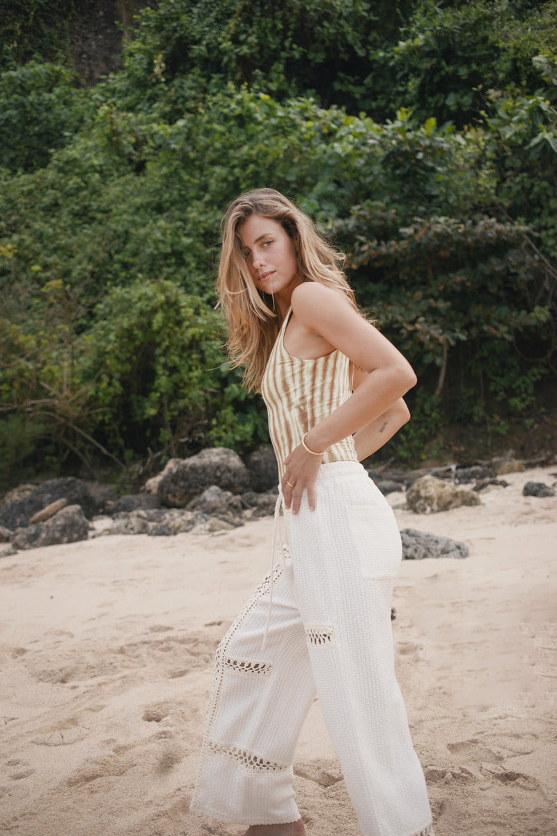 Woman in a sleeveless top and white pants standing on a beach with greenery in the background