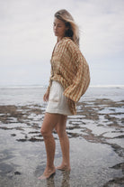 Woman standing on a rocky beach wearing a patterned shawl and white shorts.