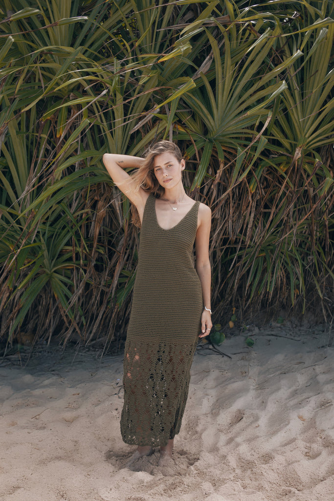 Woman in a long green dress standing on a sandy beach with tropical plants in the background