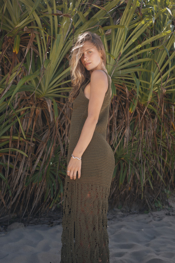 Woman wearing a long, dark green crochet dress standing against a natural background of plants and sand.