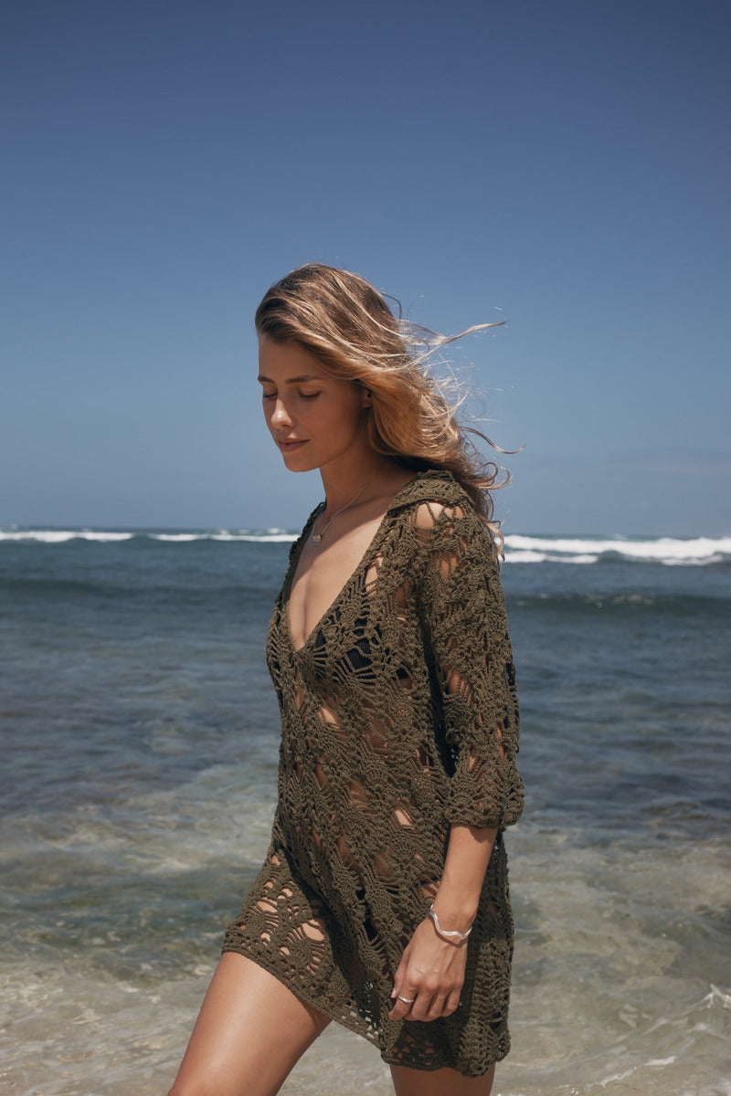Woman in a brown crochet cover-up standing on a beach with ocean waves in the background