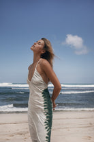 Woman in a white dress with green tie-dye pattern standing on a beach with ocean waves in the background.