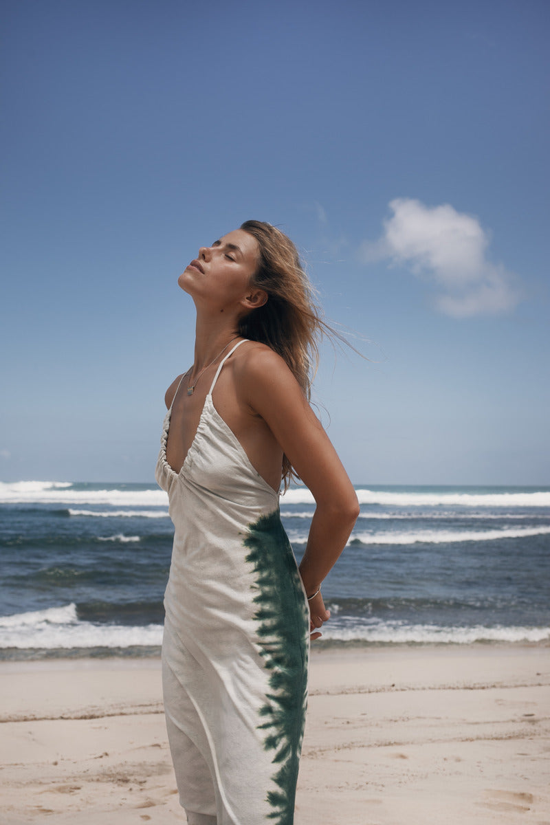 Woman in a white dress with green tie-dye pattern standing on a beach with ocean waves in the background.