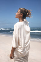 Woman in a white outfit standing on a beach with waves and clear sky.