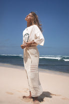 Woman in a white crochet maxi skirt standing on a beach with blue sky and ocean.