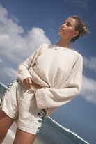 Woman in a beige top and tie dye short standing on a beach with a blue sky and clouds in the background