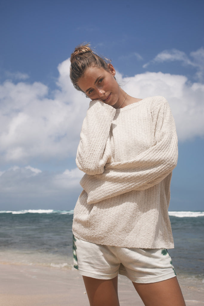 Woman wearing a cream sweater and white shorts on a beach with blue sky and ocean.