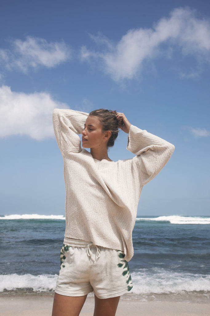 Woman in a beige sweater and shorts standing on a beach with ocean waves and blue sky.