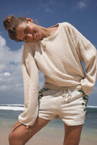Woman in a beige sweater and white shorts standing on a beach with ocean and sky in the background