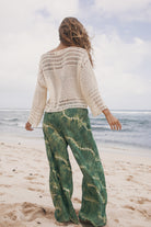 Person wearing a white crochet top and green tie-dye pants on a beach.