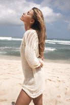 Woman in a white crochet mini dress on a beach with ocean waves in the background