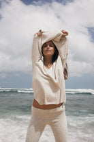 Woman in a beige outfit standing on a beach with ocean waves and cloudy sky.