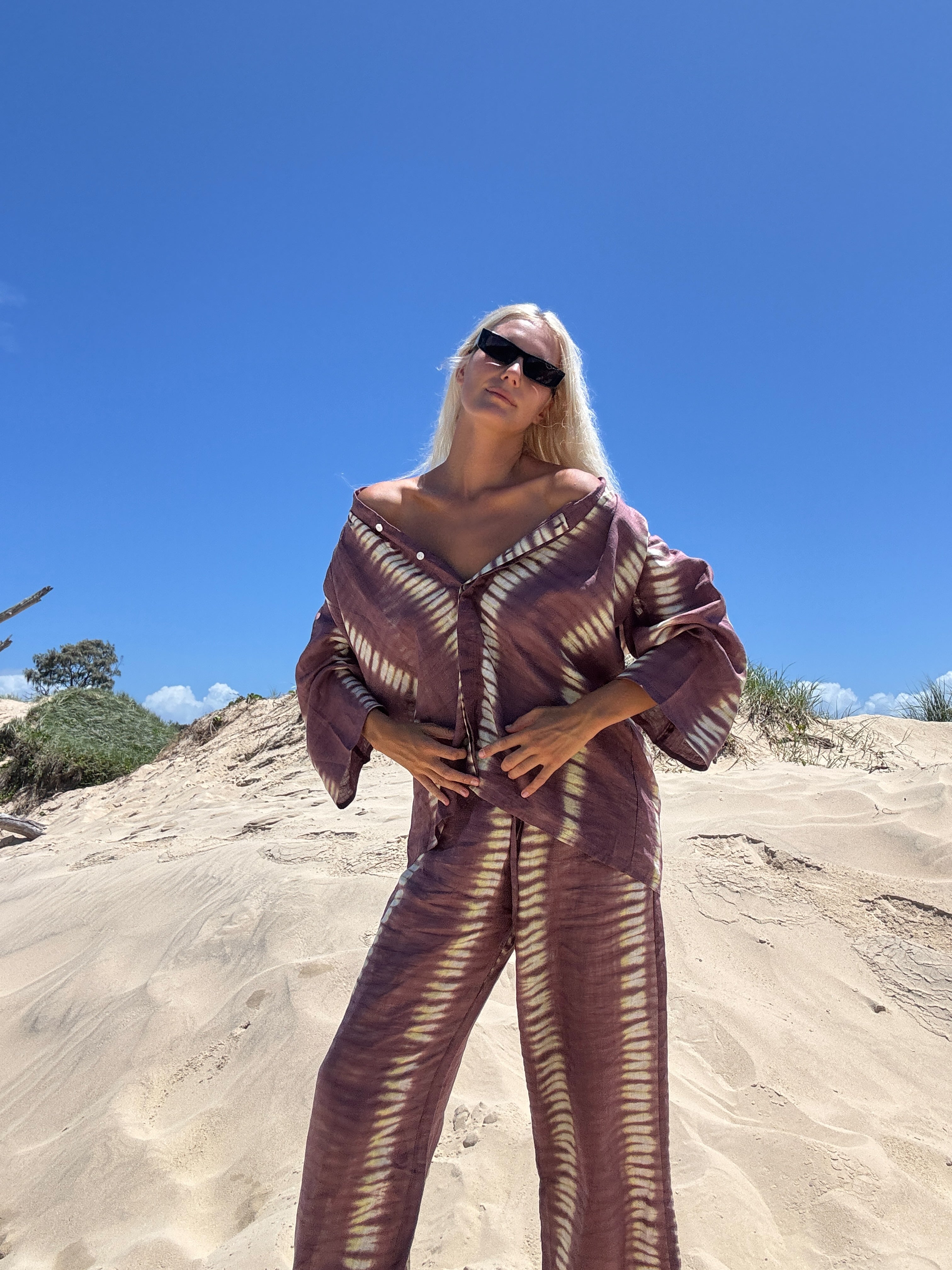 A person standing on a sandy surface wearing a deep mauve tie-dye shirt with wide sleeves and a V-neckline, paired with matching pants, sunglasses, and a handbag.